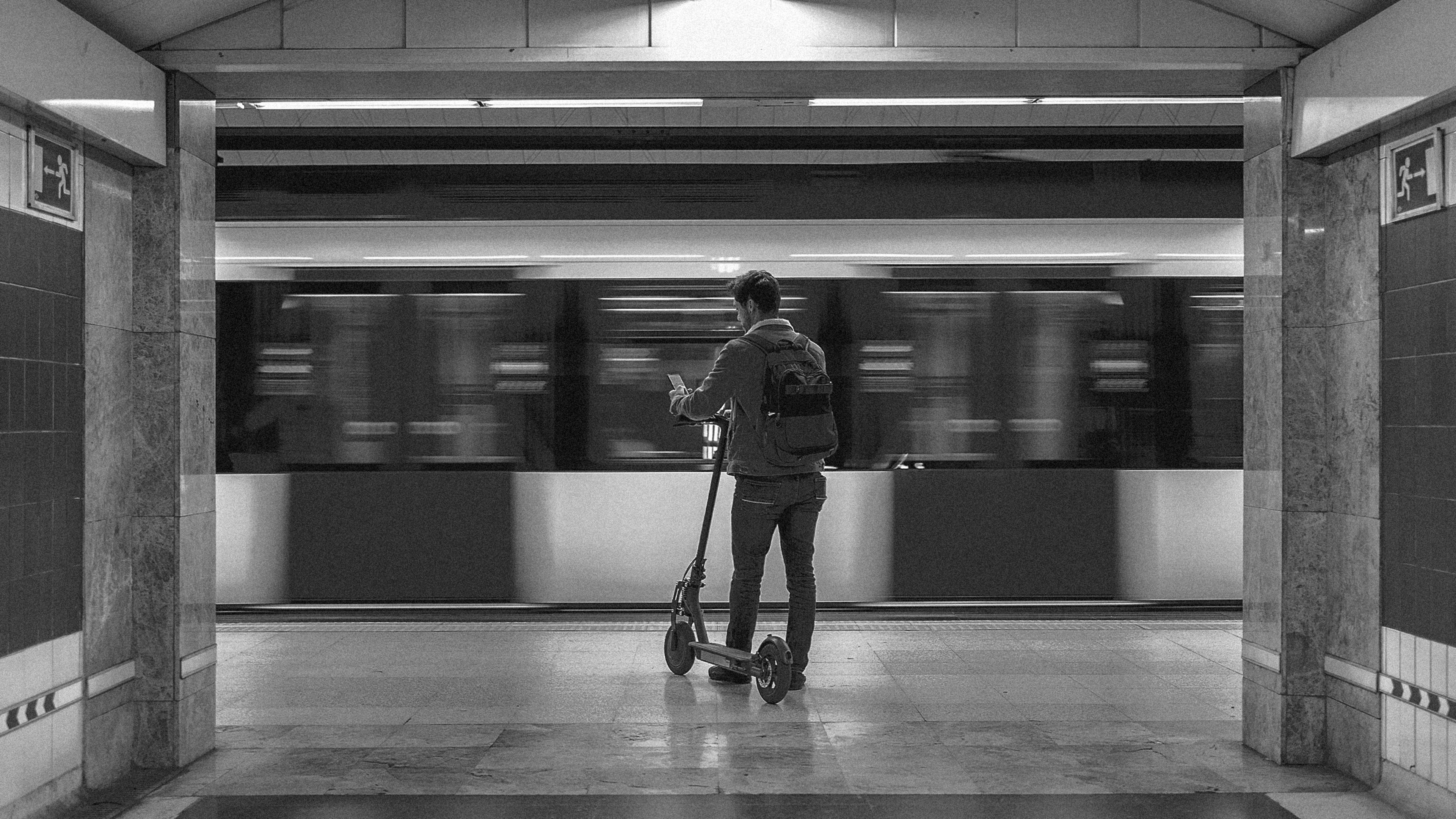 A black and white photo of a man with an electric scooter waiting for a train to stop in a subway station.