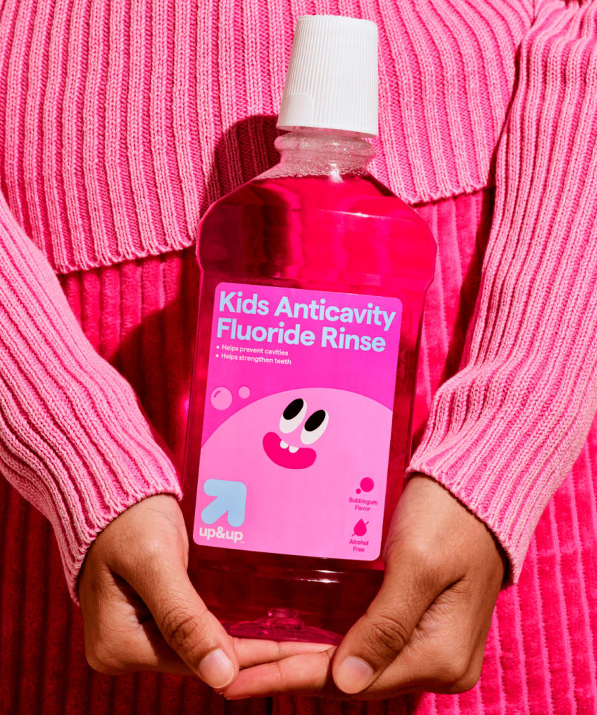 A photo two hands holding a pink package of Kids Anticavity Flouride Rinse in front of a pink outfit.