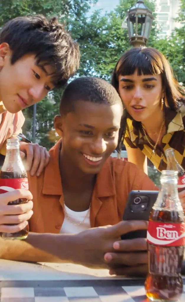A photograph of three young people looking at a phone and holding Coca Cola bottles.