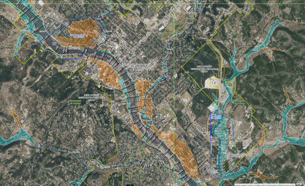 Map of Kerrville, Texas, overlaid with flood risk zones. The Guadalupe River runs centrally in purple. Blue and tan shaded areas indicate flood risks, overlaid on a detailed street grid. Roads, buildings, and green spaces are visible, showing how flood zones intersect with developed and natural areas.