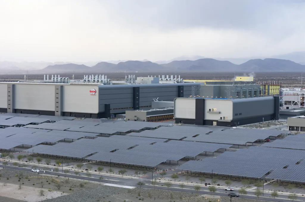 A large industrial facility in an arid region, featuring multiple buildings. The most prominent building displays the 'Intel' logo. In the foreground, rows of solar panels are visible. The buildings have rooftop structures resembling cooling towers or ventilation systems. In the background, mountains rise under a cloudy sky.