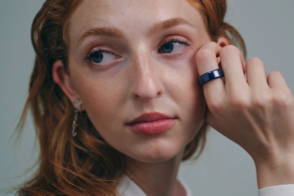 Una fotografía de cerca de una modelo sosteniendo su mano derecha sobre su rostro mientras usa un anillo Oura de cerámica azul.