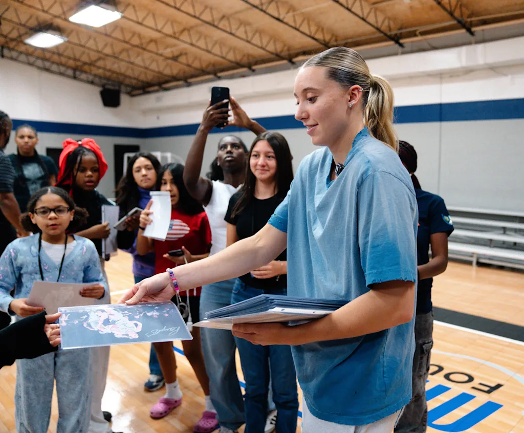 A photo of Paige Bueckers passing out signed photos of herself to young fans on a basketball court.