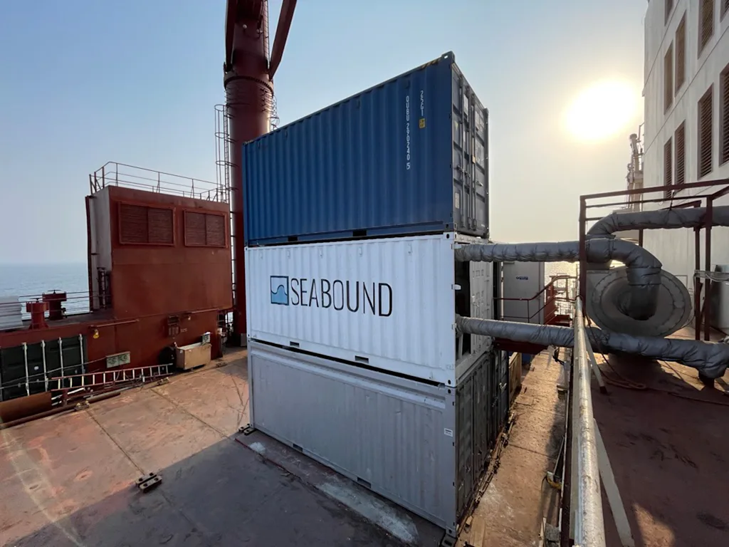 Een witte container met het Seabound-logo erop staat op het dek van een vrachtschip tussen andere zeecontainers, maar heeft twee grote pijpen die in en uit lopen en die met het schip zijn verbonden.