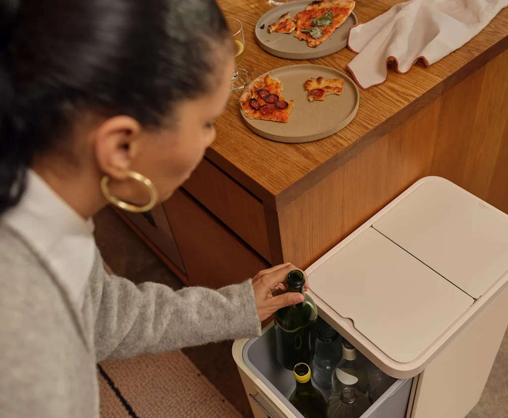 A photo of a person putting an empty wine bottle in an off-white Caraway recycling bin.