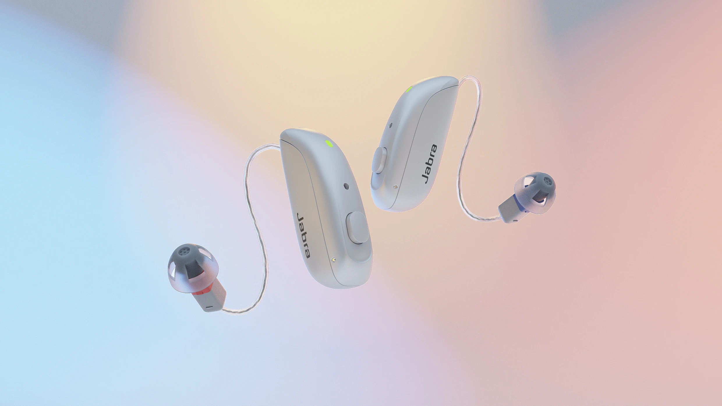 This image showcases three Jabra hearing aids against a soft, pastel-colored background that blends hues of blue, pink, and peach, creating a calming and modern aesthetic. The hearing aids are presented in a floating arrangement, seemingly suspended in mid-air, allowing for a clear view of their design and features. Two of the devices are behind-the-ear (BTE) models, one in a light beige color and the other in a white finish, both displaying the 'Jabra' logo prominently on their casing. A thin, transparent tubing connects each BTE device to a custom-molded earpiece, which is a dark grey color. The third hearing aid is a receiver-in-canal (RIC) model, also in a light beige, with a very thin wire connecting it to a smaller, darker grey earpiece. The devices are angled slightly, giving a dynamic feel to the composition. The lighting is soft and diffused, highlighting the smooth curves and subtle textures of the hearing aids. The overall impression is one of technological sophistication, discreet design, and a focus on improving quality of life through advanced hearing solutions. The background is intentionally blurred to keep the focus entirely on the products themselves, emphasizing their sleek and modern appearance.